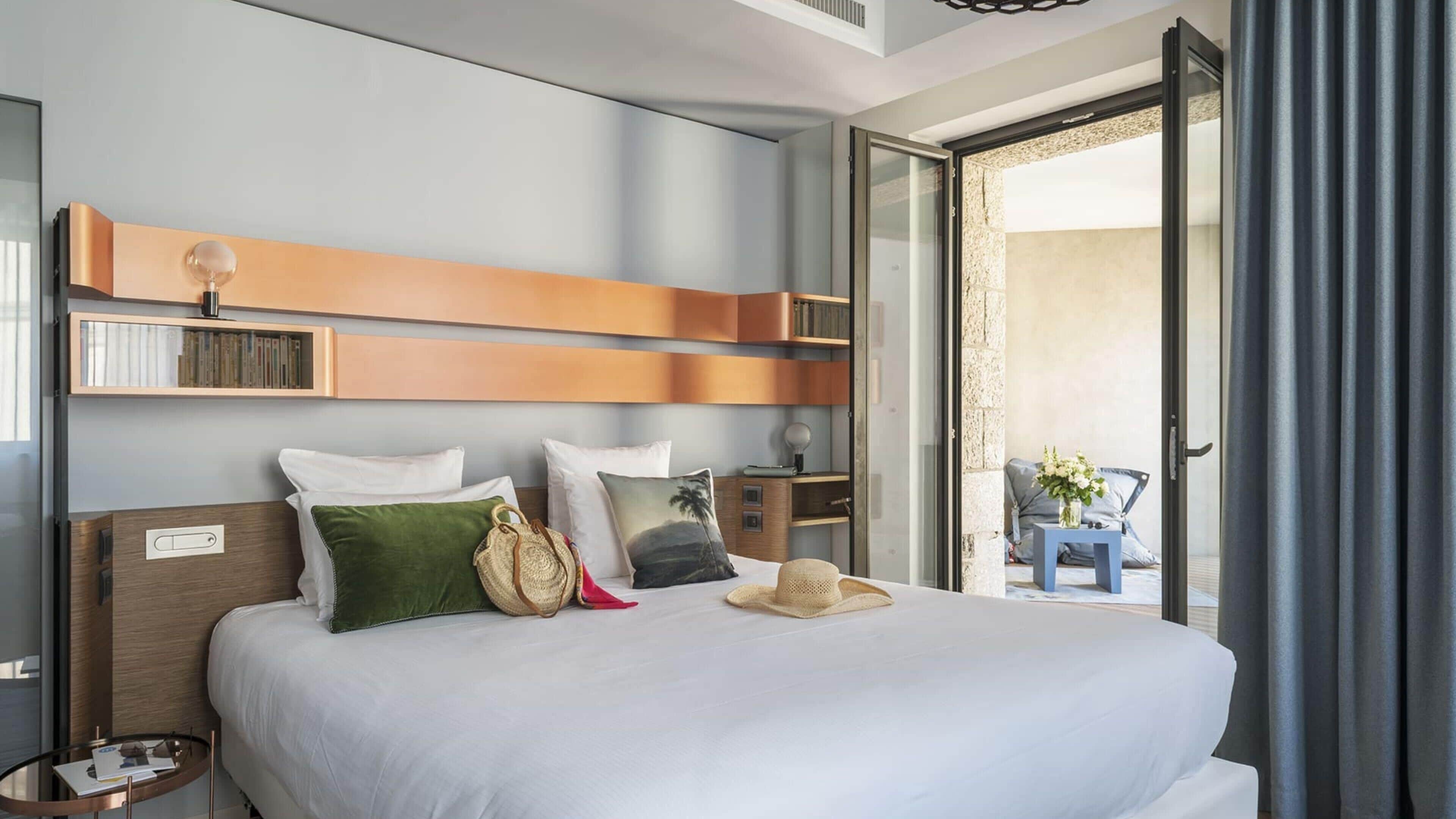 A hotel room with a bed, pillows, a bedside table, a nightstand, a wall-mounted shelf, and a view through a glass door.