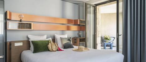 Una habitación de hotel con una cama, almohadas, una mesita de noche y una estantería en la pared, además de vistas a través de un ventanal.