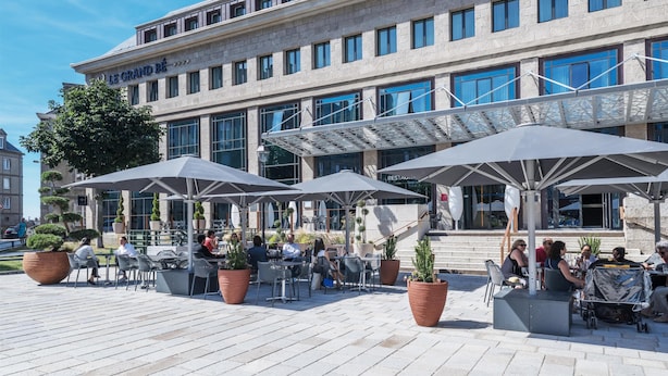 An outdoor café with tables and umbrellas, a building with a sign reading 'Le Grand Bé', and potted plants. - Golden Tulip Saint Malo - Le Grand Bé (Saint-Malo)
