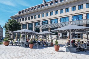 An outdoor café with tables and umbrellas, a building with a sign reading 'Le Grand Bé', and potted plants. - Golden Tulip Saint Malo - Le Grand Bé (Saint-Malo)