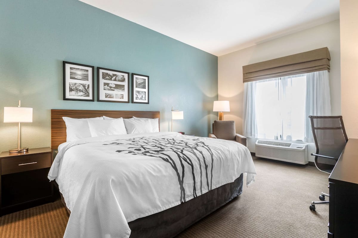 Ein Hotelzimmer mit einem großen Bett, zwei Nachttischen mit Lampen, einem Sessel, einem Schreibtisch und einem großen Fenster mit Vorhängen.
