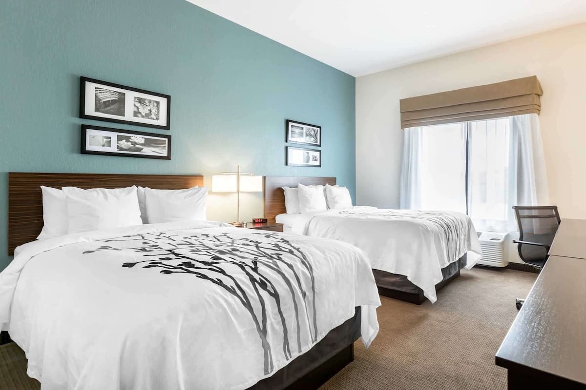 Ein Hotelzimmer mit zwei Betten, einem Schreibtisch und einem Stuhl. An der Wand hängen Bilder in Rahmen und ein Fenster mit Vorhängen.