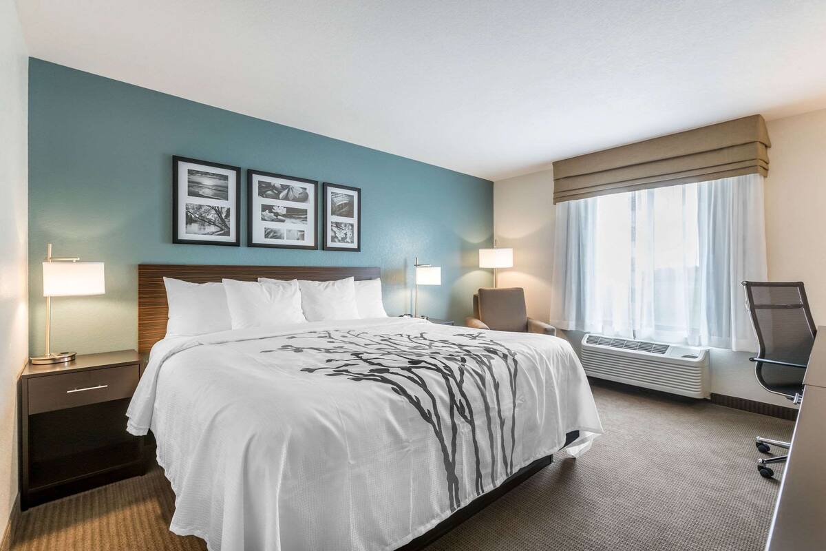 Ein Hotelzimmer mit Bett, Nachttischlampen, einem Sessel, einem Schreibtisch und einem fenster mit Vorhängen.