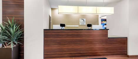 Un espace d’accueil avec un comptoir de réception en bois, deux haut-parleurs noirs, une enseigne indiquant « Sleep Inn by Choice Hotels », une plante en pot et un tapis noir portant le nom de l’hôtel.