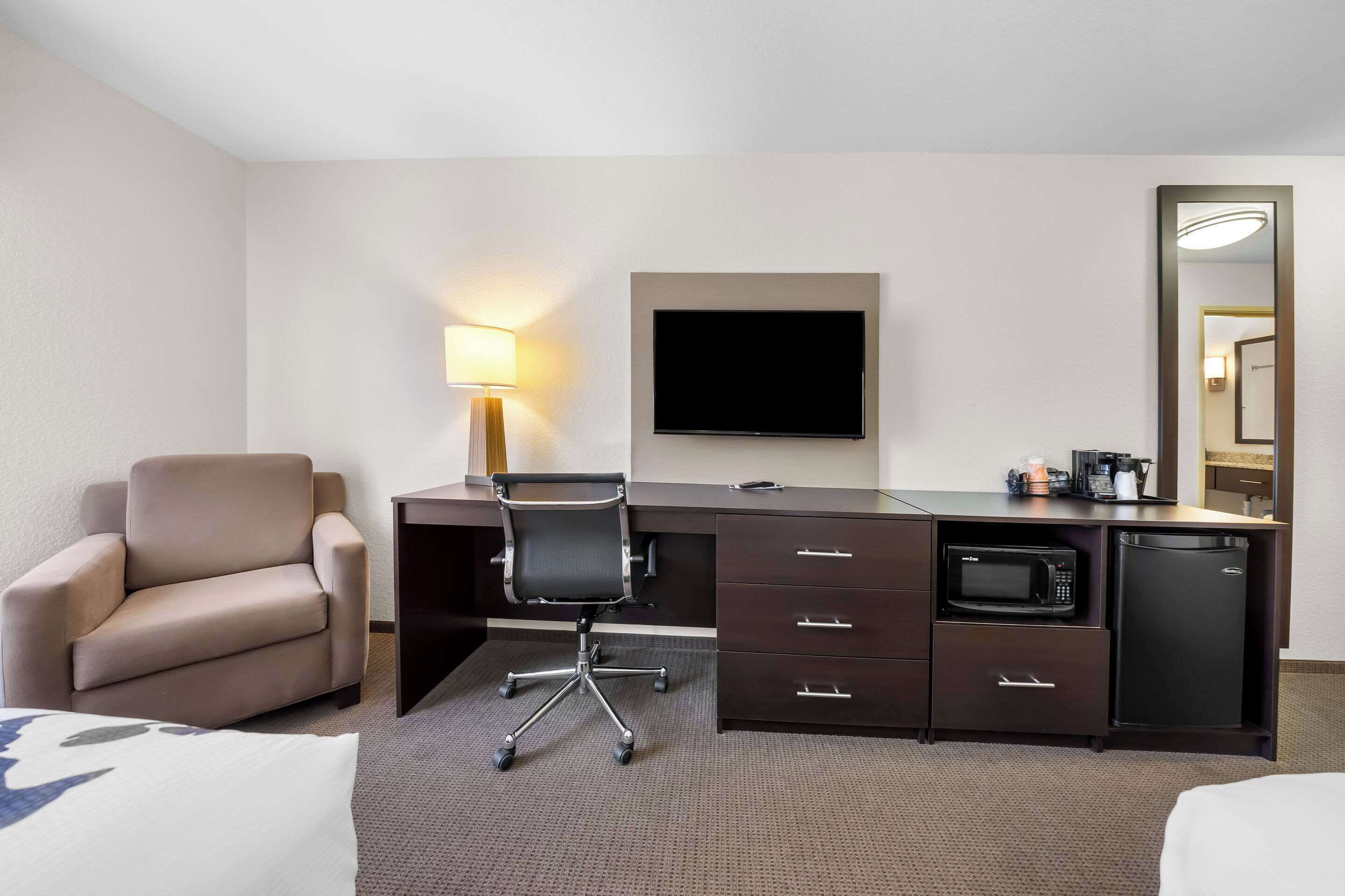A hotel room with a bed, a desk with a chair, a flat-screen TV, a microwave, a refrigerator, a lamp, and a mirror.