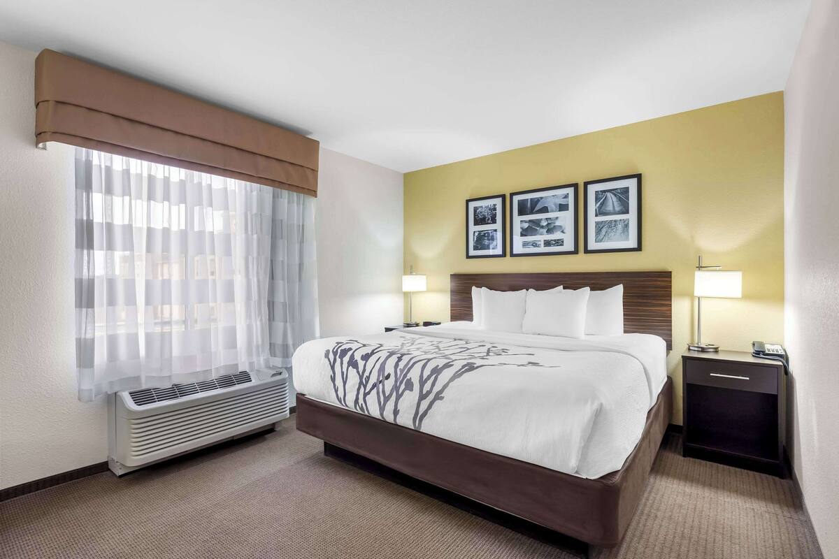 A hotel room with a large bed, a bedside table with a lamp, a wall-mounted air conditioner, and framed pictures on the wall.