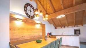 A wooden dining table with chairs, a clock, pendant lights, and a kitchen in the background. - Luxury apartment in Brixen (Brixen im Thale)