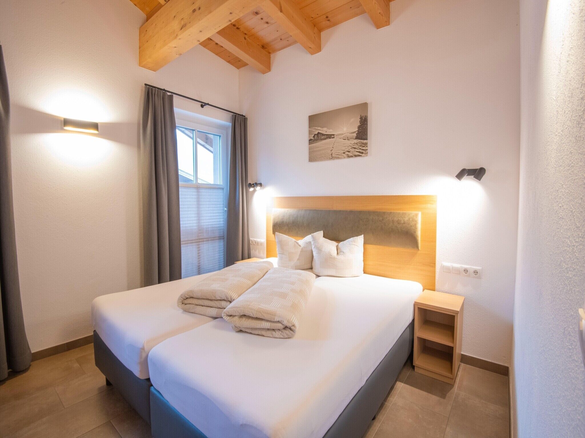 A hotel room with a double bed, a headboard with integrated lighting, a bedside table, and a window with curtains.