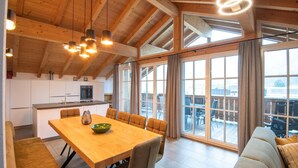 A spacious dining area with a wooden table and chairs, a kitchen in the background, and large windows offering a view of the outdoors. - Luxury apartment in Brixen (Brixen im Thale)