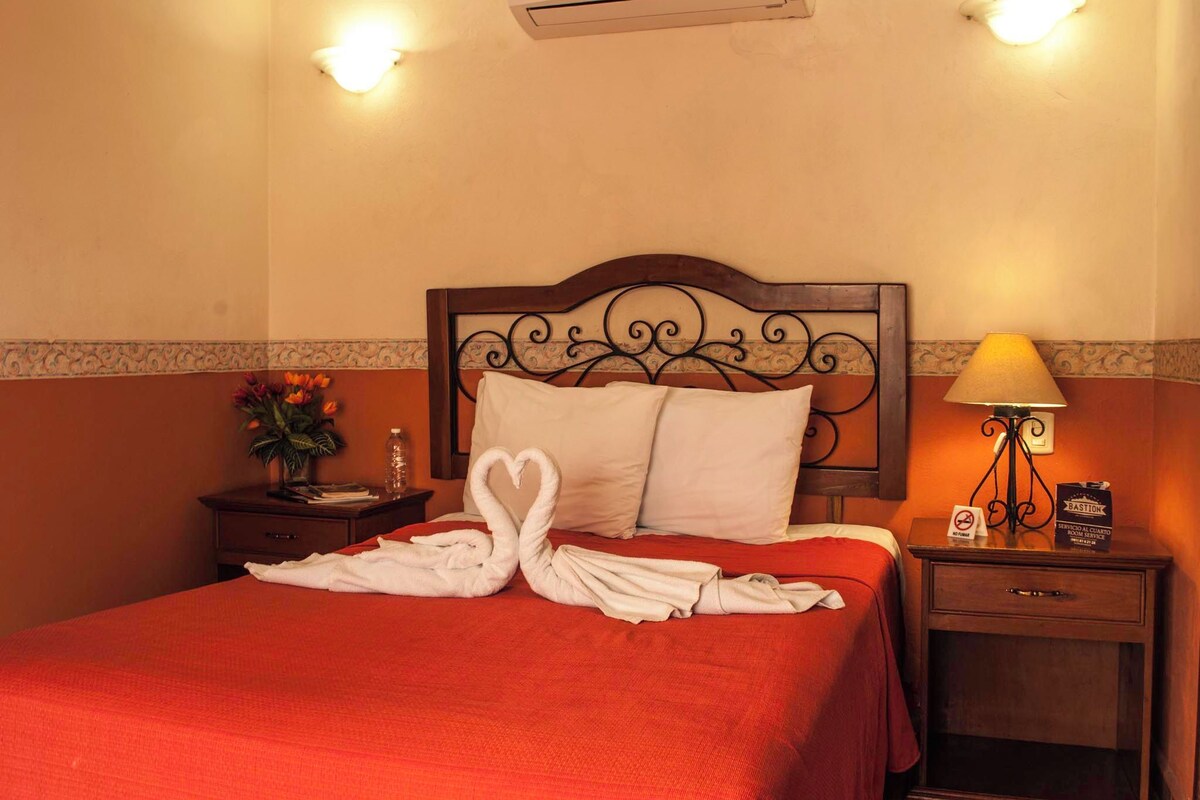 A hotel room with a red bedspread, a wooden headboard, a nightstand with a lamp, and a wall-mounted air conditioner.
