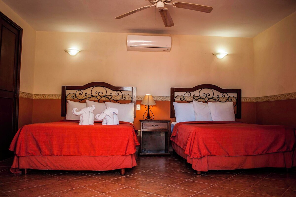 A hotel room with two beds, each with a red bedspread and white pillows, a nightstand with a lamp, and an air conditioning unit on the wall.