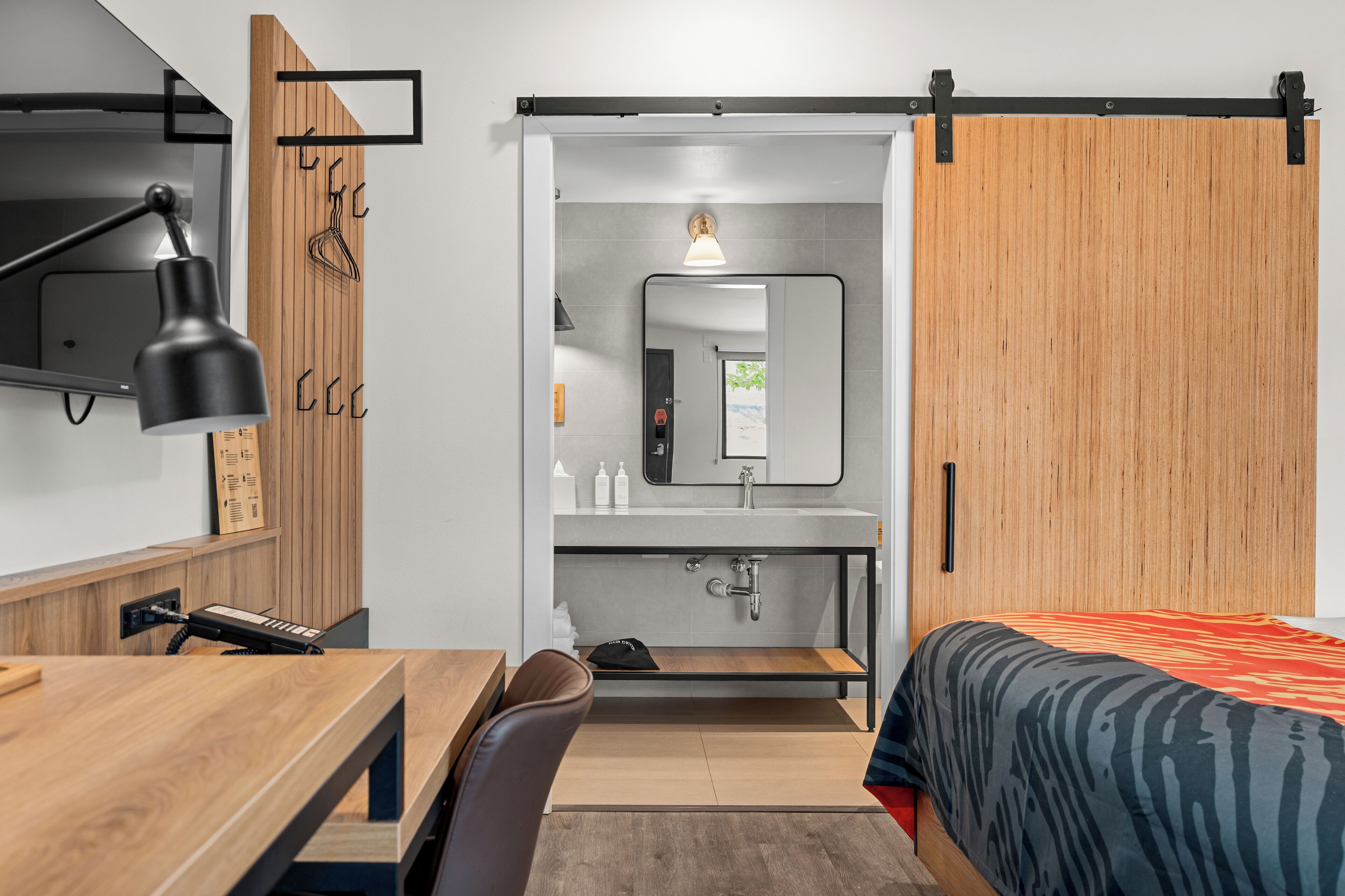 A modern hotel room with a wooden desk, a black desk lamp, a bed with a zebra print bedspread, a sliding door closet, and a bathroom with a mirror and sink.