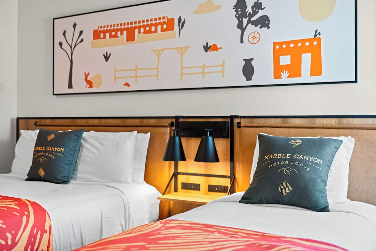 A hotel room with two beds, each with a pillow featuring 'Marble Canyon Motor Lodge' text. There is a wooden headboard, a nightstand with two black lamps, and a wall art depicting a desert scene.