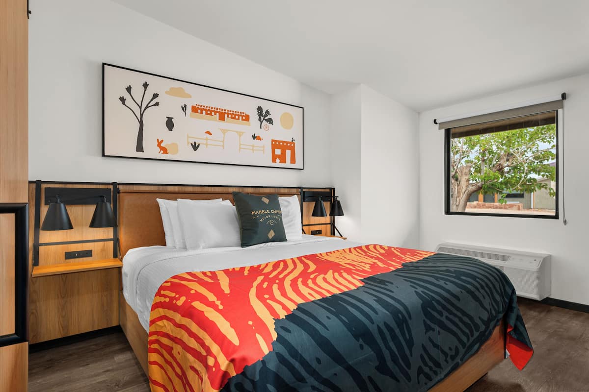 A hotel room with a bed, a nightstand, a wall-mounted lamp, a window with a view, and a framed picture on the wall.