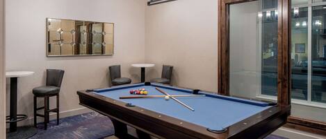 Une table de billard recouverte d’un drap bleu, entourée de quatre chaises, et un lustre au-dessus.