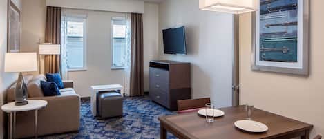 Habitación de hotel con sofá, una mesita, una mesa de comedor con platos y vasos, televisión y un cuadro en la pared.