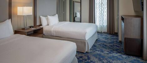 A hotel room with two beds, a desk with a lamp, a mirror, a wardrobe, and a patterned carpet.