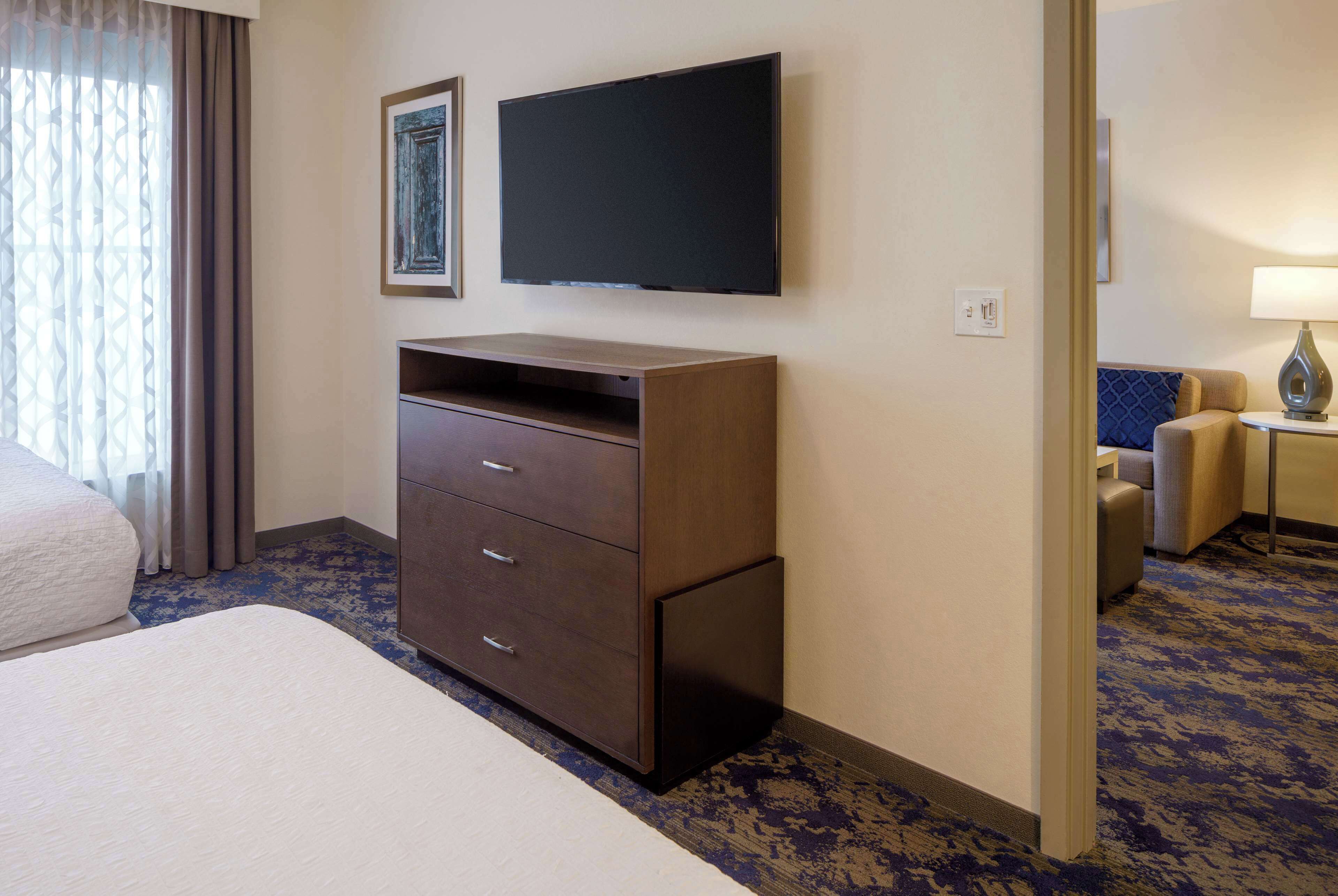 A hotel room with a large bed, a wooden dresser, a flat-screen TV, and a seating area with a lamp.