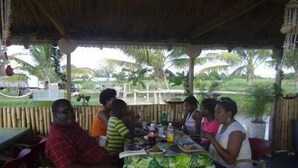 A group of people dining at an outdoor restaurant with a thatched roof, surrounded by palm trees and greenery. - Kiskeya Guest House (Léogâne)