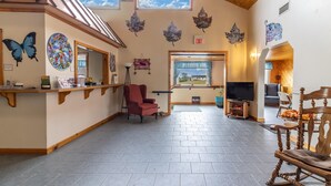 A spacious lobby with a high ceiling, wooden beams, a reception desk, a red armchair, a television, and a view of the outdoors through large windows. - Gateway Inn (Medway)