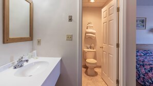 A bathroom with a white sink, mirror, and toilet. There is a door leading to another room with a bed. - Gateway Inn (Medway)