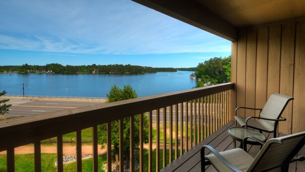 A wooden deck with two chairs and a small table, overlooking a lake with a road and greenery in the background. - The Pointe Hotel and Suites (Minocqua)