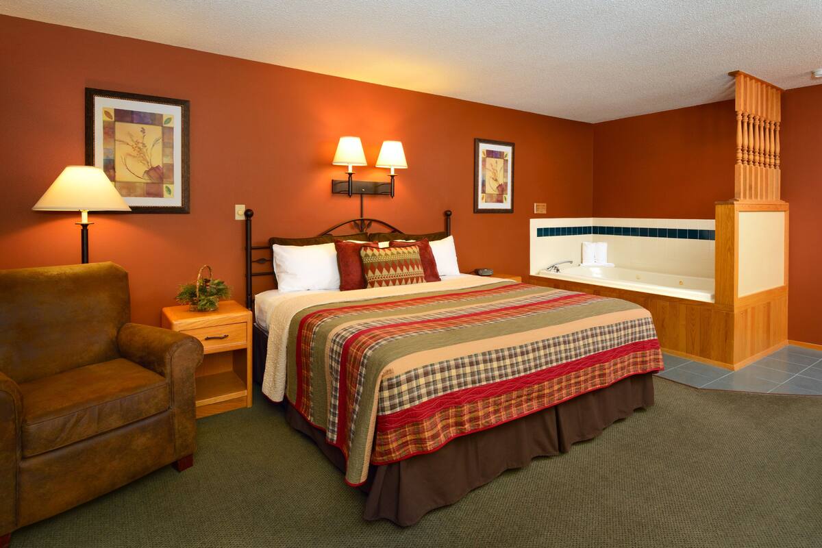 A hotel room with a bed, a nightstand, a lamp, a chair, a bathroom with a bathtub, and a wooden column.