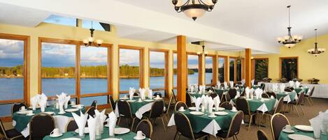 Un comedor con mesas dispuestas para el servicio, amplias ventanas con vistas a un lago y candelabros.