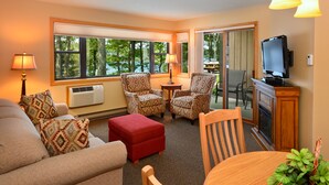 A cozy living room with a sofa, two patterned armchairs, a red ottoman, a wooden dining table with a plant, and a television on a wooden cabinet. - The Pointe Hotel and Suites (Minocqua)