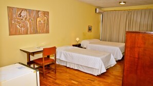 A hotel room with two beds, a desk, a chair, a refrigerator, and a painting on the wall. - Hotel Estoril (Pelotas)