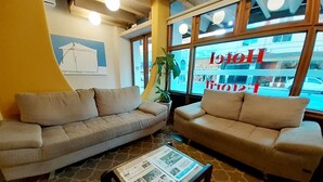 A hotel lobby with two sofas, a coffee table with a newspaper, and a large window with the hotel's name. - Hotel Estoril (Pelotas)