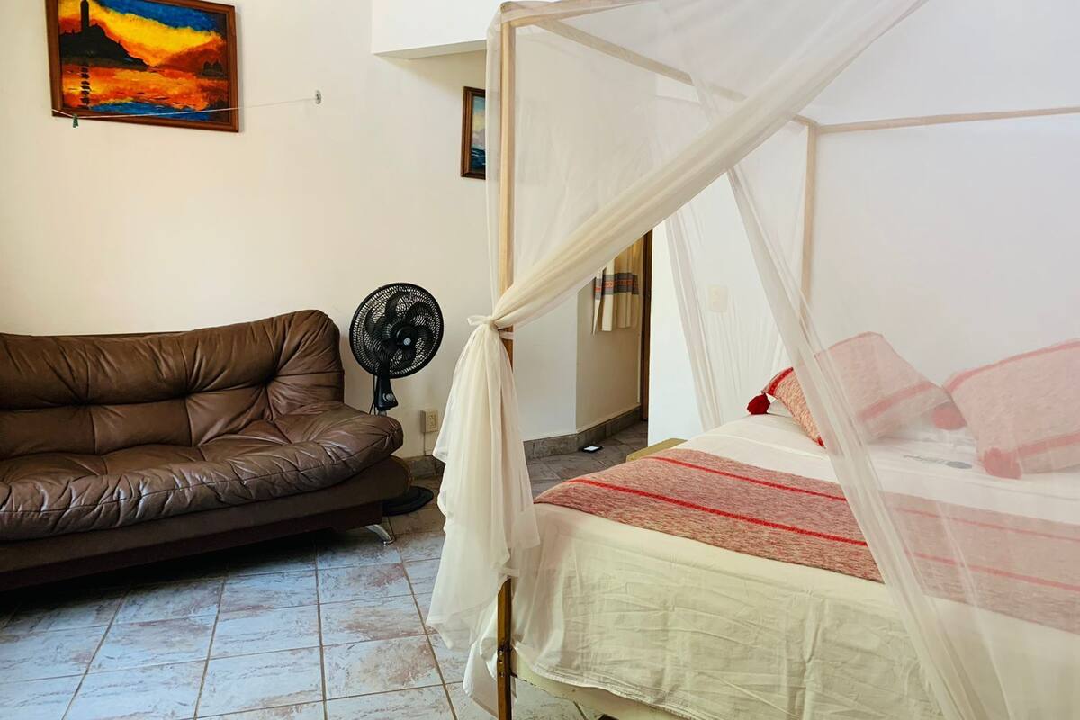 Une chambre à coucher avec un lit sous un dais, un canapé en cuir marron, un ventilateur sur pied et un tableau accroché au mur.