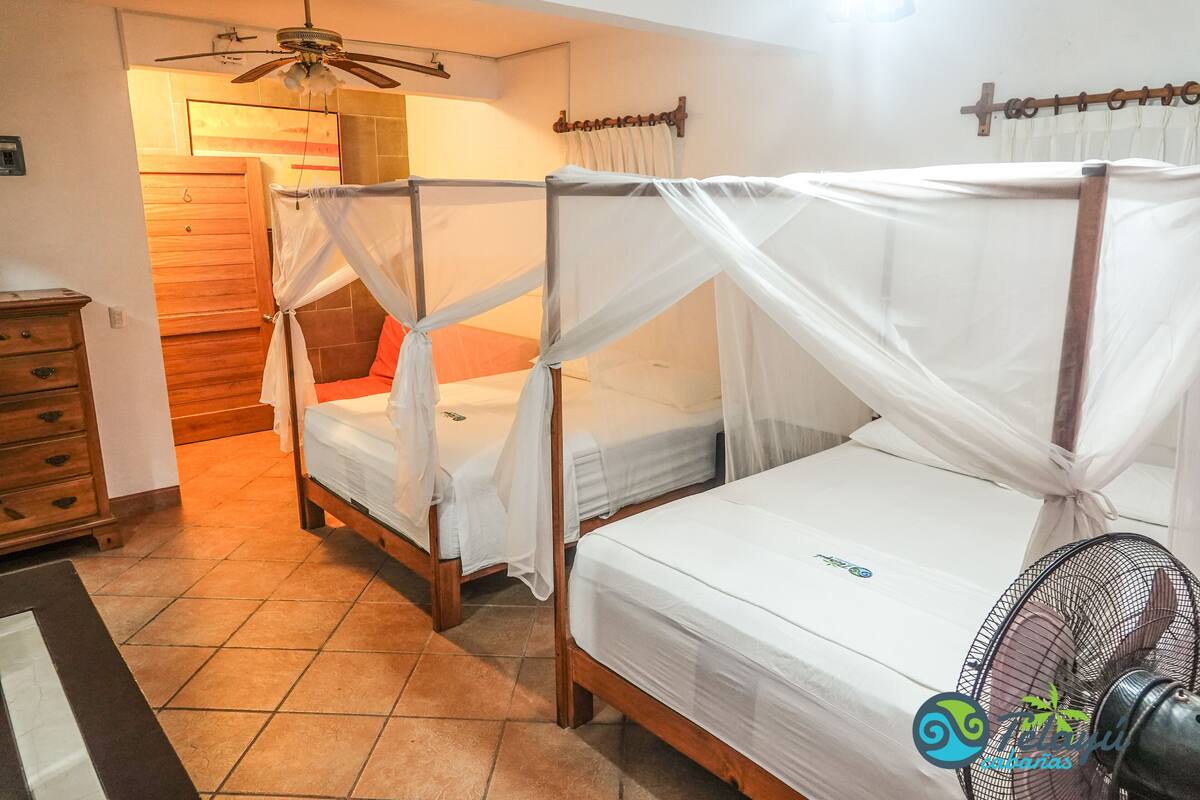 Une chambre à coucher avec deux lits en bois, chacun surmonté d’un baldaquin, une commode en bois et un ventilateur de plafond.