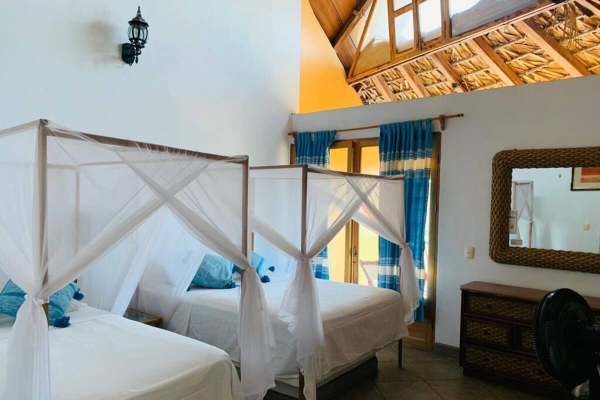 Une chambre avec deux lits, chacun surmonté d’un baldaquin blanc. Le plafond est en bois et présente un décor de chaume, et la pièce est équipée d’un ventilateur mural et d’un miroir.
