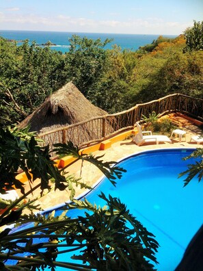 A pool area with a thatched roof structure, lounge chairs, and a view of the ocean. - Cabañas Telayú (Santa María Tonameca)