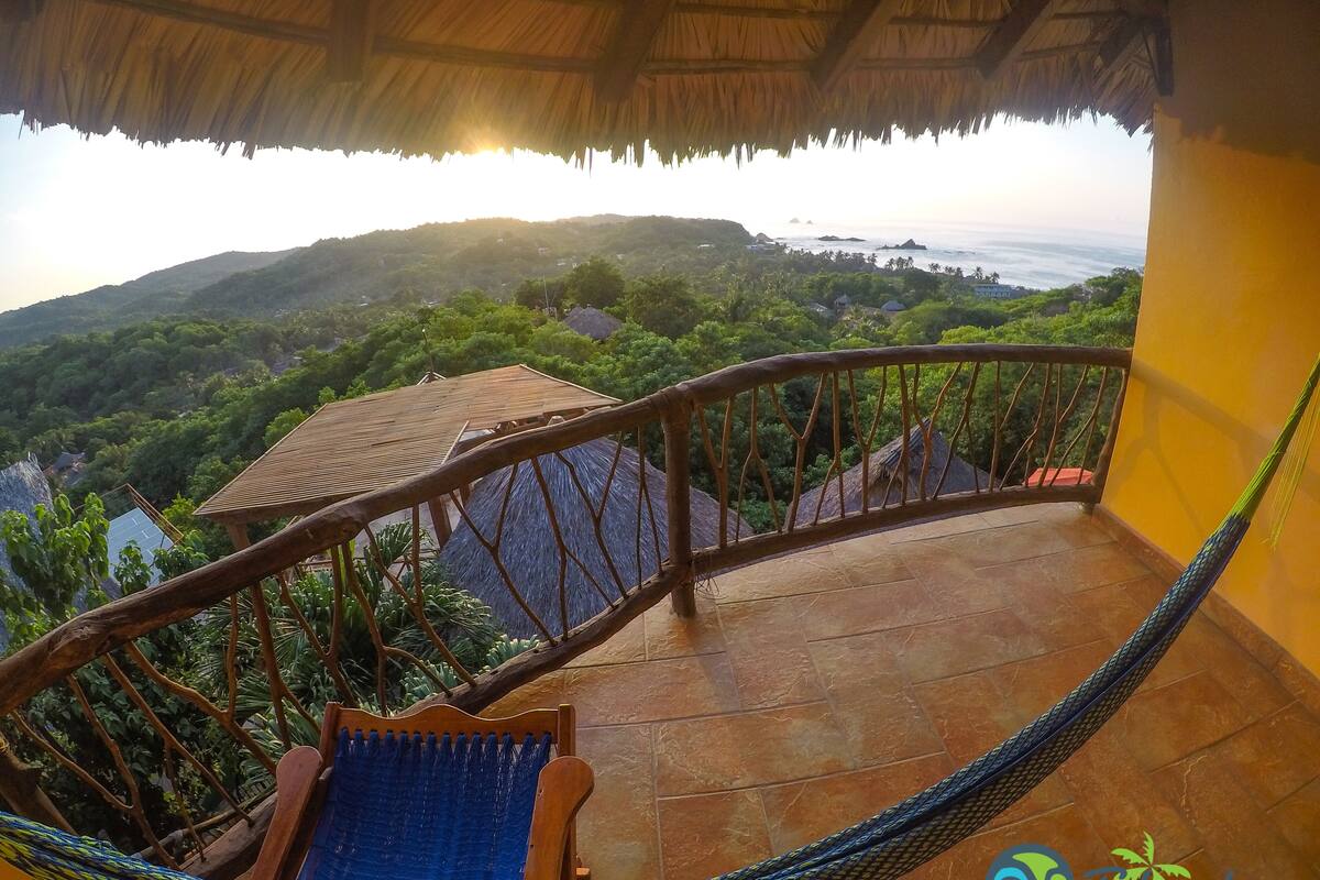 Un balcon doté d’un hamac, de balustrades en bois et offrant une vue sur un paysage luxuriant et l’océan.