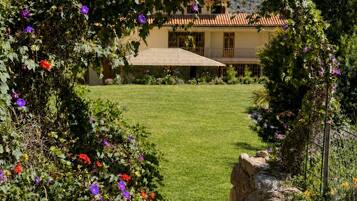 Un chemin de pierres mène à une maison dont le toit est recouvert de tuiles, et qui est entourée d’un jardin où poussent des fleurs violettes, avec une montagne en arrière-plan.