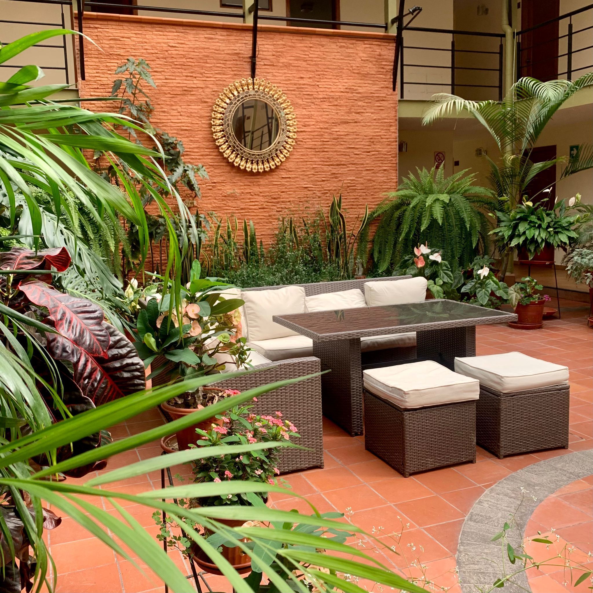 An outdoor seating area with wicker furniture and a glass-top table, surrounded by various potted plants and a decorative mirror on a brick wall.