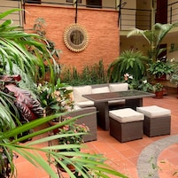 An outdoor seating area with wicker furniture and a glass-top table, surrounded by various potted plants and a decorative mirror on a brick wall.
