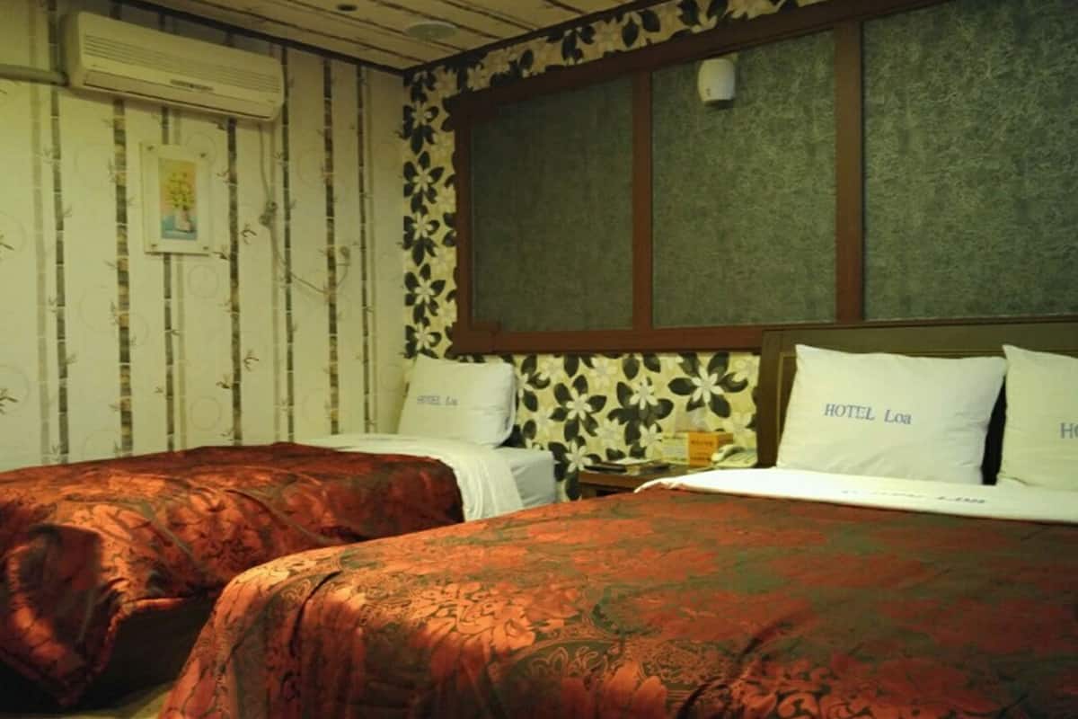 A room with two beds, each with a floral-patterned bedspread and pillows labeled "HOTEL Loa". The room has a wooden ceiling and walls with a decorative panel.