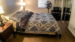 Une chambre à coucher avec un lit, une lampe de chevet, un ventilateur sur pied, une télévision et un miroir.