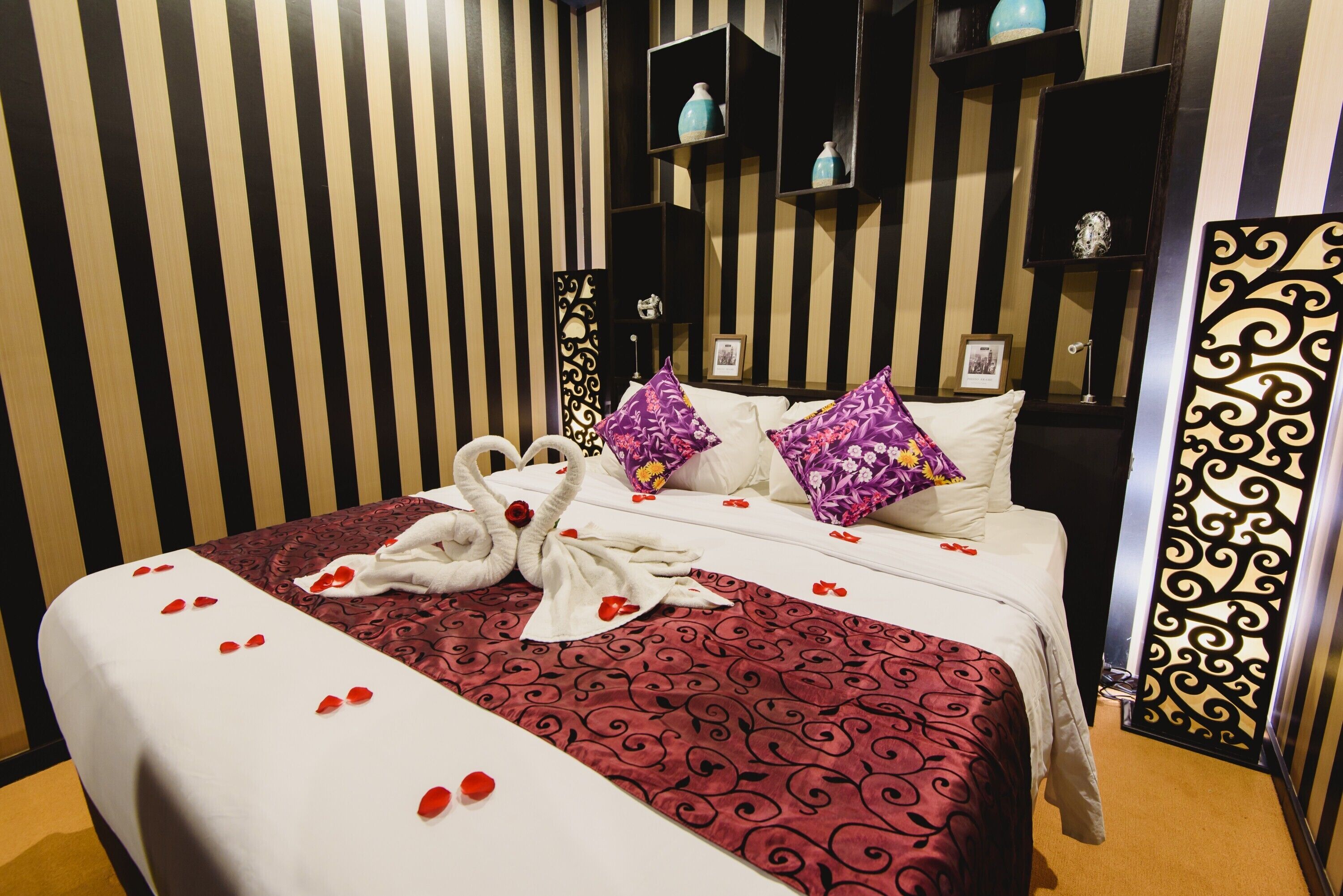 A hotel room with a striped wall, a bed with a red patterned bedspread, white pillows, and a decorative towel arrangement.