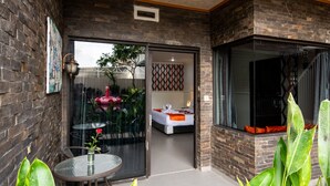 A modern hotel room with a glass door leading to a balcony, a bed with a red and white patterned headboard, and a dining area with a table and chairs. - Hotel L'Amore Bali (Seminyak)