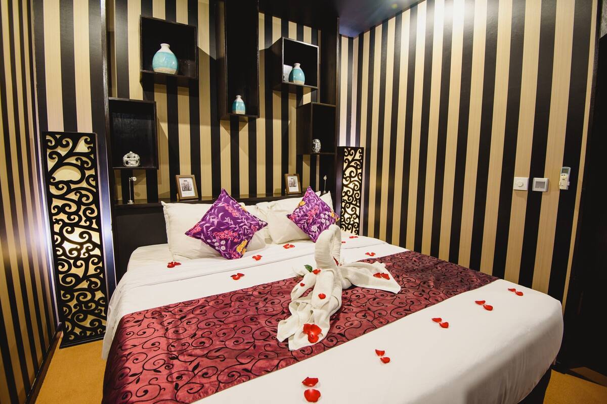 A hotel room with striped wallpaper, a bed with a red and white patterned bedspread, and decorative pillows.