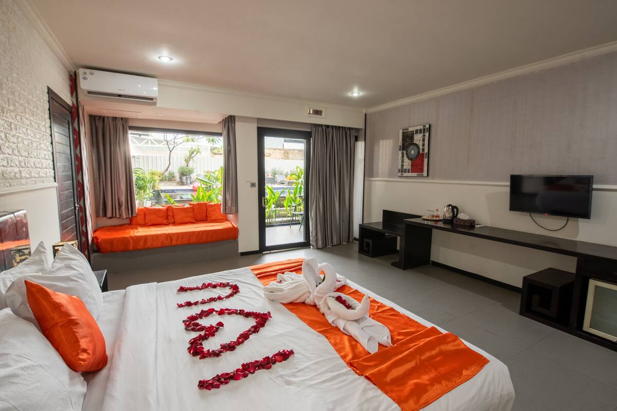 A hotel room with a bed, a desk, a TV, and a balcony with plants.