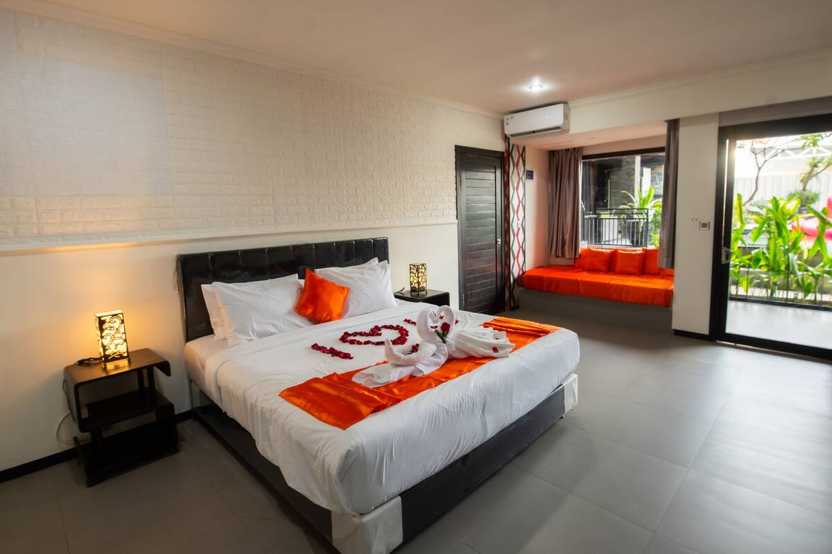 A modern hotel room with a large bed, a nightstand, a balcony with orange cushions, and a view of greenery.
