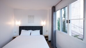 Ein modernes Hotelzimmer mit einem großen Bett, zwei Nachttischen, einer Wandleuchte und einem fensterbelaubten Fenster.