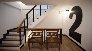 Eine Treppe mit Holzhinweisen und schwarzen Geländern, die zu einem Podest mit zwei Holzsesseln und einer großen schwarzen Zahl '2' an der Wand führt.