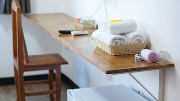 A compact room with a wooden desk, a chair, a small white refrigerator, a wall-mounted TV, and a basket of folded towels.