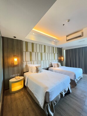 A hotel room with two beds, a wooden floor, a modern headboard, and a small bedside table with a lamp.